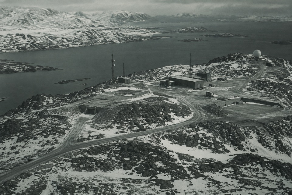 Desaturated aerial reconstruction of a remote northern military facility with few buildings, antennas and access roads.